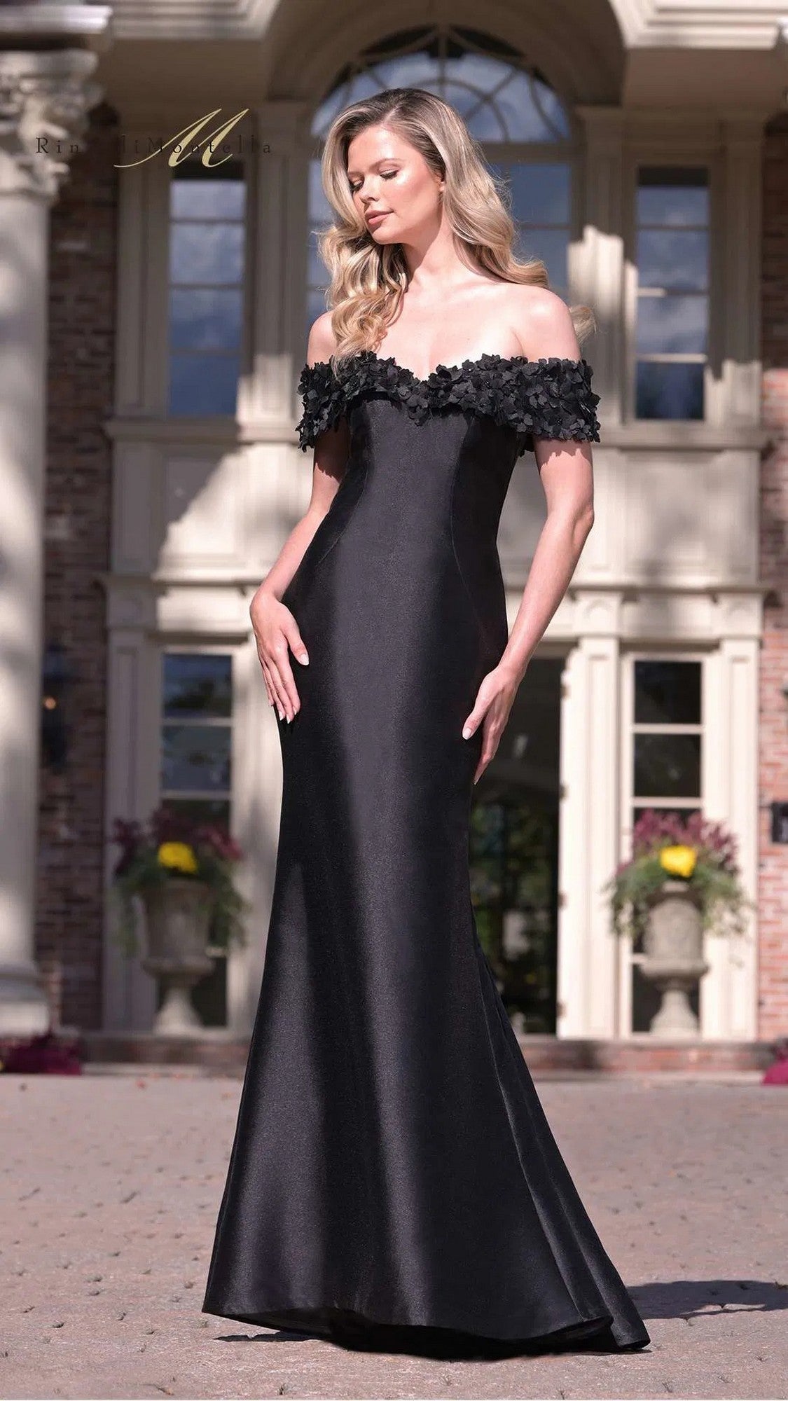 Outdoors, a woman wears the Rina Di Montella RD3254—a long, formal, black mermaid evening dress with off-the-shoulder floral-detailed bodice.